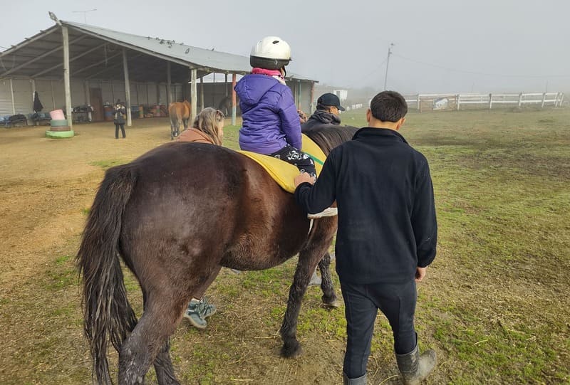Un niño con discapacidad está montando un caballo, con un joven y una mujer a su lado, ayudándolo y apoyándolo